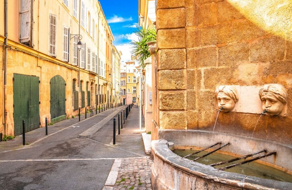 Quiet historic street in Aix-en-Provence with a traditional stone wall fountain and pastel buildings under blue sky